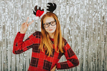 Christmas portrait of cute little girl against silver background, wearing red pullover and reindeer earsの写真素材