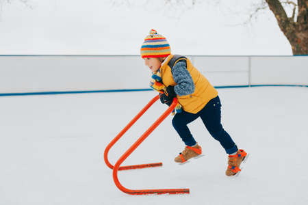 Happy funny little boy practicing with support on skating rink, wearing bright hat and yellow jacket. Winter activities for childrenの写真素材