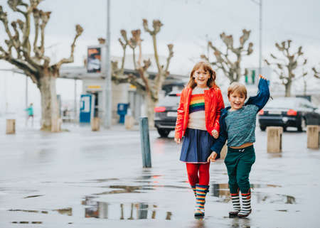 Two funny kids, little brother and sister playing together in a city under the rain in early spring. Chidren wearing rain jackets and bootsの写真素材
