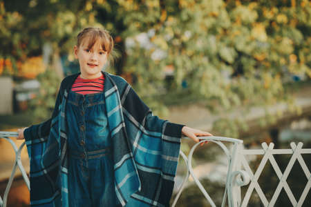Fashion portrait of pretty little girl wearing blue plaid poncho, denim overalls, standing on the bridge on a nice sunny dayの写真素材