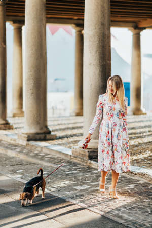 Young blond woman walking her dog on city street, wearing beautiful dressの写真素材