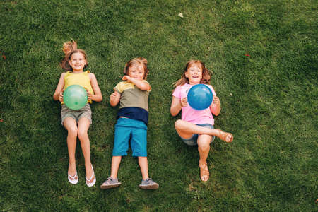 Happy children having fun outdoors. Kids playing in summer park. Little boy and two girls lying on green fresh grass, holding balloonsの写真素材
