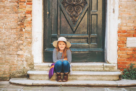 Outdoor portrait of pretty kid girl walking through old italian streets, wearing frock, denim jeans and neutral straw hatの写真素材