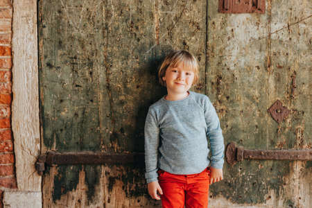 Outdoor portrait of handsome little boy wearing red chinos and light blue pullover, stylish kid posing against vintage old wall. Fashion for small childrenの写真素材