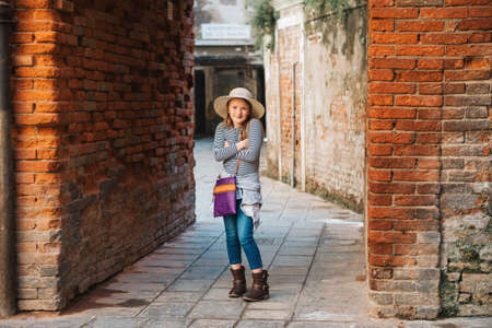 Outdoor portrait of pretty kid girl walking through old italian streets, wearing frock, denim jeans and neutral straw hatの写真素材
