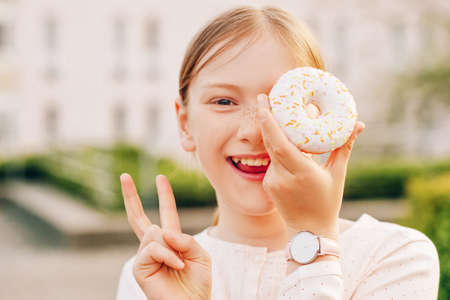 Close up portrait of funny little girl playing with donut, wearing watch, showing peace or victory sign with fingersの写真素材