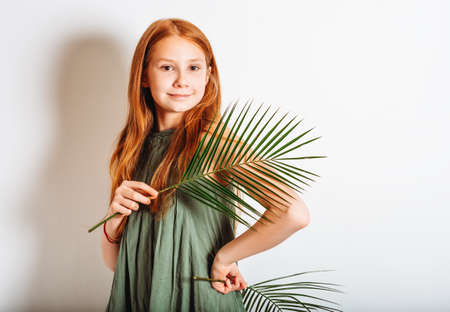 Studio shot of adorable red-haired preteen kid girl, wearing khaki dress, playing with exotic green palm leavesの写真素材