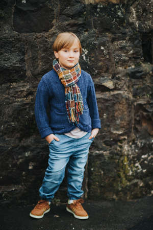 Outdoor portrait of handsome little boy wearing denim jeans, blue knitted jacket and brown shoes, stylish kid posing against dark wall. Fashion for small childrenの写真素材