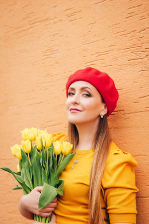 Outdoor spring portrait of young beautiful woman, wearing yellow top and red beret, holding bouquet of fresh tulip flowers, posing against orange wallの写真素材