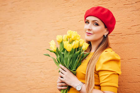 Outdoor spring portrait of young beautiful woman, wearing yellow top and red beret, holding bouquet...