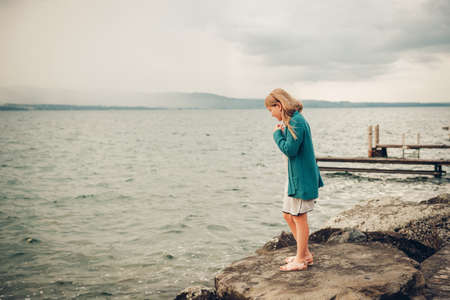 Outdoor portrait of adorable little girl playing next to lake, wearing white dress and green cardigan jacketの写真素材