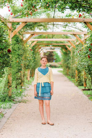 Outdoor portrait of adorable preteen 10 year old girl in beautiful rose garden, wearing blue dress and yellow jacketの写真素材