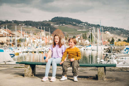 Two funny kids, little brother and sister, playing together outside by the lake, resting on the bench in small Lutry port, Lausanne area, Switzerlandの写真素材