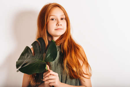Studio shot of adorable red-haired preteen kid girl, wearing khaki dress, playing with exotic green Monstera leafの写真素材