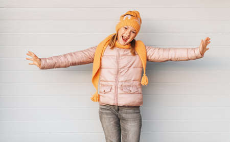 Expressive child posing against white background, wearing soft pink padded jacket and set of orange knitted hat and scarf. Winter fashion for childrenの写真素材