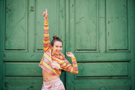 Young expressive woman posing next to green old door, wearing short pink and yellow stripe sweatshirt, pointing upの写真素材