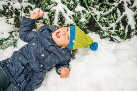 Happy little boy playint with snow outside, wearing warm winter jacket and knitted hat, lying in snow, top viewの写真素材