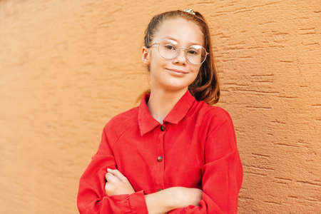 Outdoor portrait of cute preteen girl wearing red dress and eyeglasses, posing against orange wallの写真素材