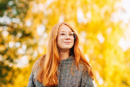 Outdoor close up portrait of beautiful red-haired preteen girl wearing check coat and eyeglasses