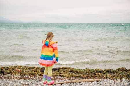 Cute little girl playing by the lake on a windy day, wearing colorful parka and rain boots. Image taken on Lake Geneva, Lausanne, Switzerland. Back viewの写真素材