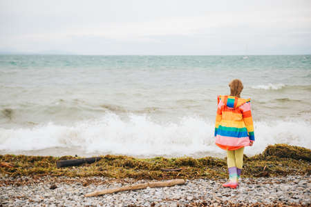 Cute little girl playing by the lake on a windy day, wearing colorful parka and rain boots. Image taken on Lake Geneva, Lausanne, Switzerland. Back viewの写真素材