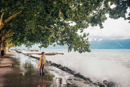 Cute little boy playing by the lake on a very windy day, watching waves, wearing yellow rain coat. Image taken on lake Geneva, Lausanne, Switzerlandの写真素材