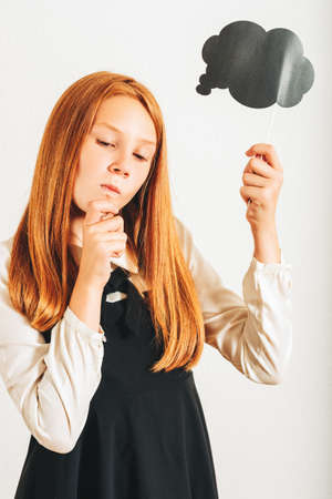 Studio shot of young red-haired kid girl holding paper thinking bubble, posing on white background