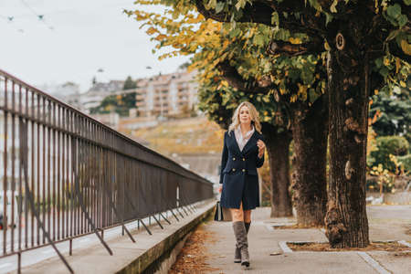 Outdoor portrait of beautiful woman wearing dark blue jacket dress, model walking down the street, holding a bagの写真素材