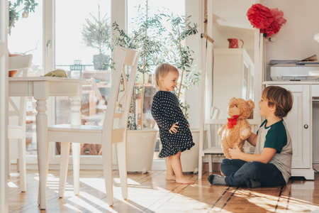 Two funny kids playing together with teddy bear at home in white living room filled with sunlightの写真素材