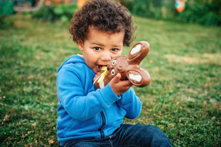 Adorable two year old african toddler boy eating chocolate bunny on backyard