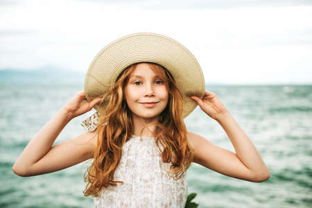 Outdoor portrait of pretty young kid girl with red hair, wearing big hat, family vacation with children by the seaの写真素材