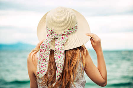 Outdoor portrait of pretty young kid girl with red hair, wearing big hat, family vacation with children by the sea. Back viewの写真素材