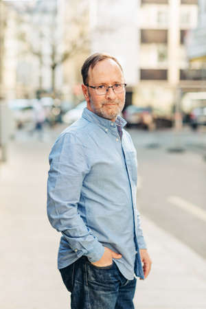 Outdoor portrait of handsome middle age man wearing long sleeve Oxford shirt, posing on city street, holding hand in jeans pocketの写真素材