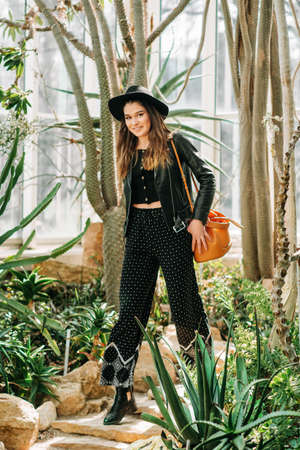 Outdoor portrait of beautiful young woman wearing black leather jacket and hat, green tropical leaves on background