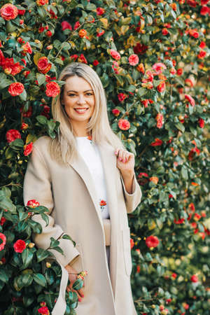 Outdoor portrait of beautiful blond woman wearing beige coat, posing in spring garden