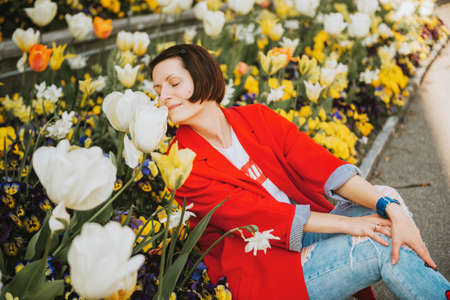 Spring portrait of pretty young woman posing against blooming tulips, wearing red jacketの写真素材