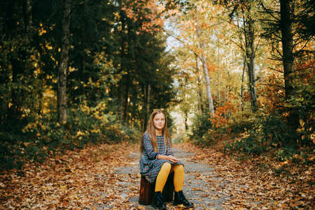 Autumn portrait of pretty young girl with old vintage suitcase, kid model posing outdoor in the forest, wearing plaid dress and yellow tightsの写真素材