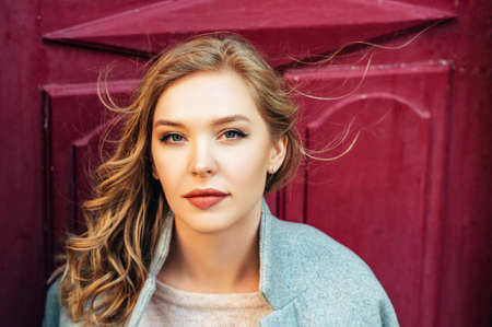 Outdoor close up portrait of beautiful woman with blond curly hair, posing next to burgundy color...
