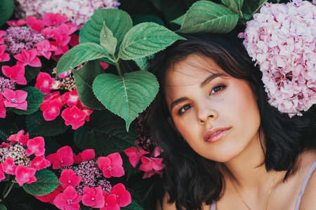 Outdoor portrait of beautiful young woman posing in pink hydrangea flowers