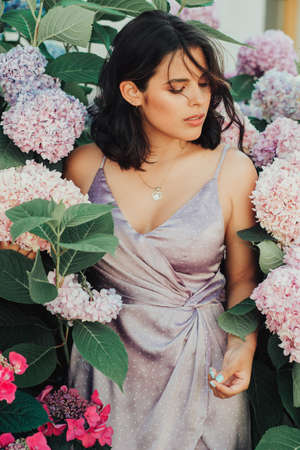 Outdoor portrait of beautiful young woman posing in pink hydrangea flowers