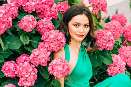 Outdoor portrait of beautiful young woman posing in pink hydrangea flowers, wearing green dressの写真素材