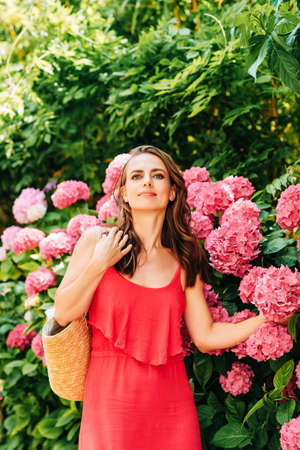 Outdoor portrait of beautiful woman posing in pink hydrangea flowers, wearing red dress