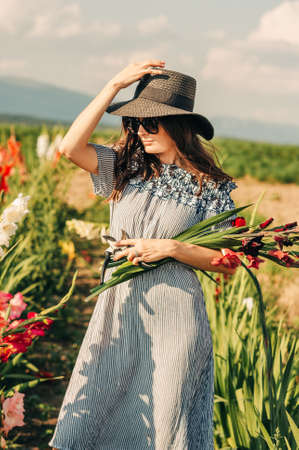 Gorgeous young woman picking flowers in a field, wearing summer dress, black straw hat and sunglassesの写真素材