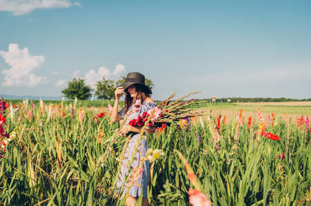 Gorgeous young woman picking flowers in a field, wearing summer dress, black straw hatの写真素材
