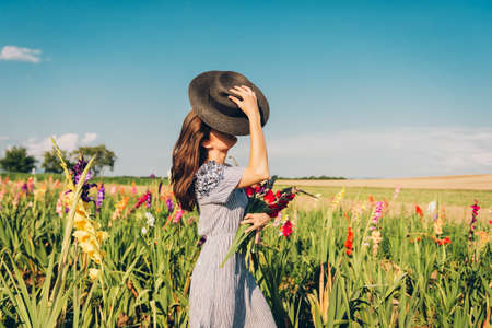 Gorgeous young woman picking flowers in a field, wearing summer dress, black straw hatの写真素材