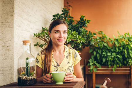 Romantic portrait of beautiful young woman wearing yellow dress, relaxing on the balcony between many green plants, holding cup of tea or coffeeの写真素材