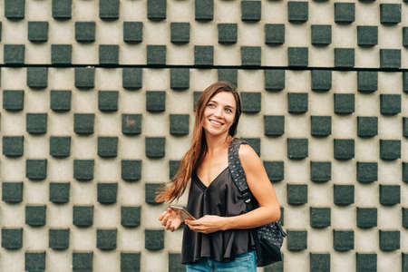Outdoor portrait of attractive young woman with long brown hair, wearing backpack, holding phoneの写真素材