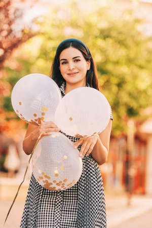 Beautiful young woman with brunette wave hair,  wearing black and white dress, holding golden balloonsの写真素材