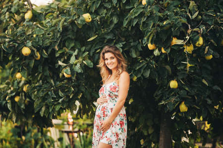 Outdoor portrait of beautiful pregnant woman wearing floral dress, posing in sunny garden