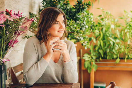 Romantic portrait of beautiful young woman wearing grey pullover, relaxing on the balcony between many green plants, holding cup of tea or coffeeの写真素材